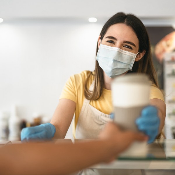 Une jeune femme portant un masque facial et des gants distribuant un café de sortie au-dessus d’un comptoir